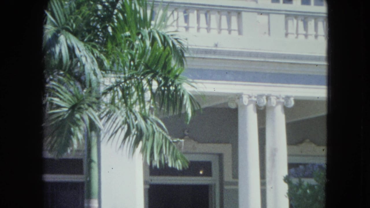 1964 San Juan home-movie view of a colonial-style building facade with white columns, balcony railings, and palm fronds.