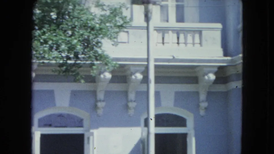 1964 San Juan home-movie frame of the building's lower facade with arched windows, balcony railing, palm trunk, and tree foliage.