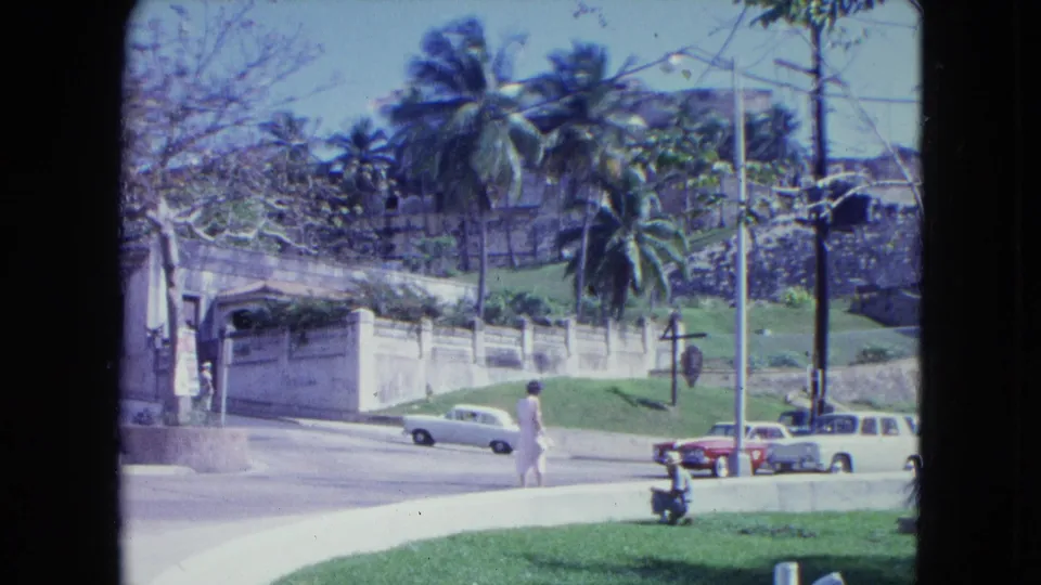 1964 San Juan street scene with a pedestrian, period cars, palm trees, a stone wall, and bright lawn in a handheld 8mm frame.