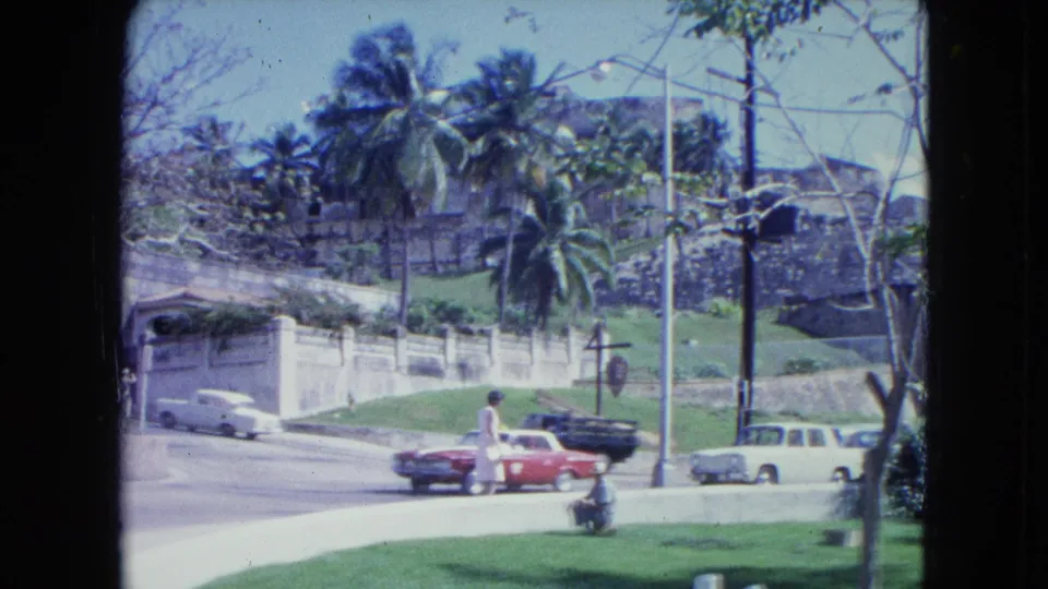 1964 San Juan frame showing period cars on a curved street beside palms, lawn, and an older stone wall, with a distant person visible.