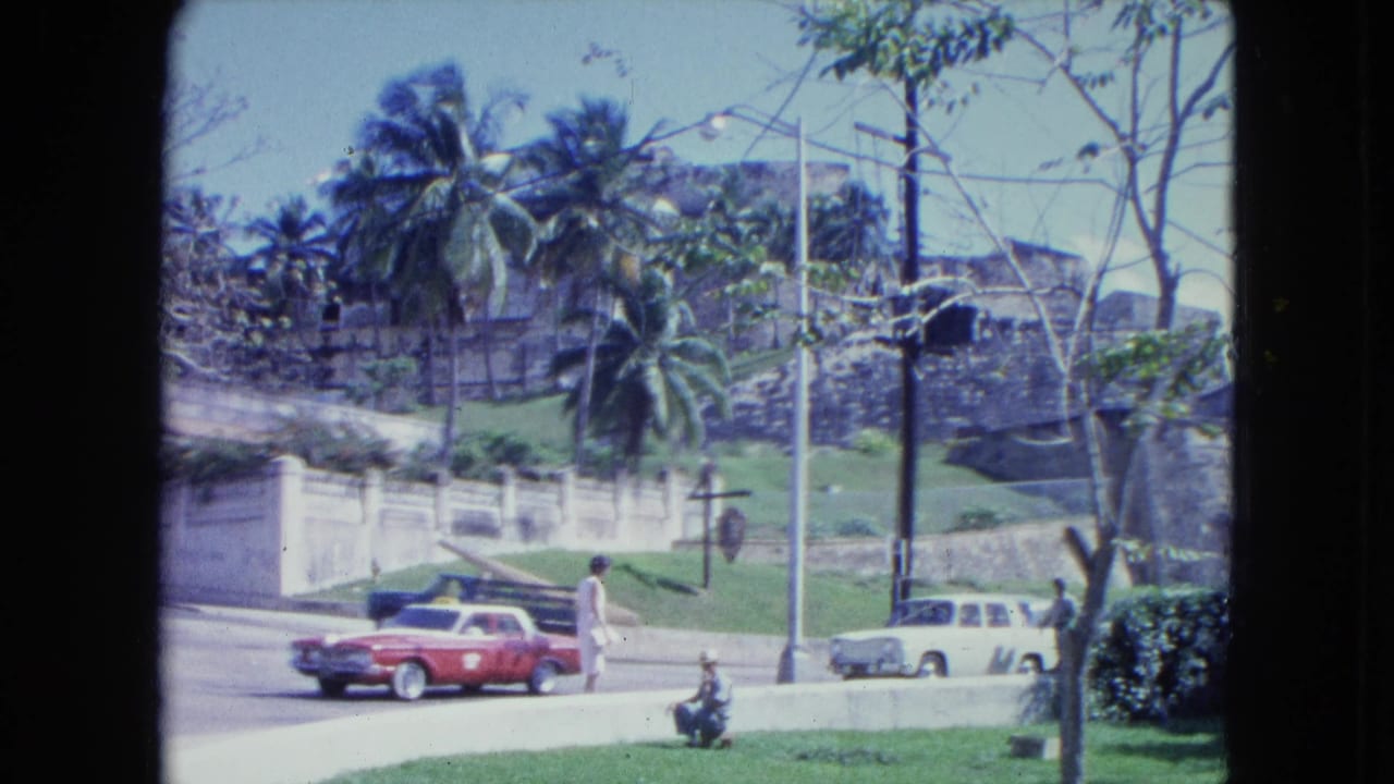 Sunny street with red car, white station wagon, pedestrians, palms, and a stone fortification beyond a grassy slope.