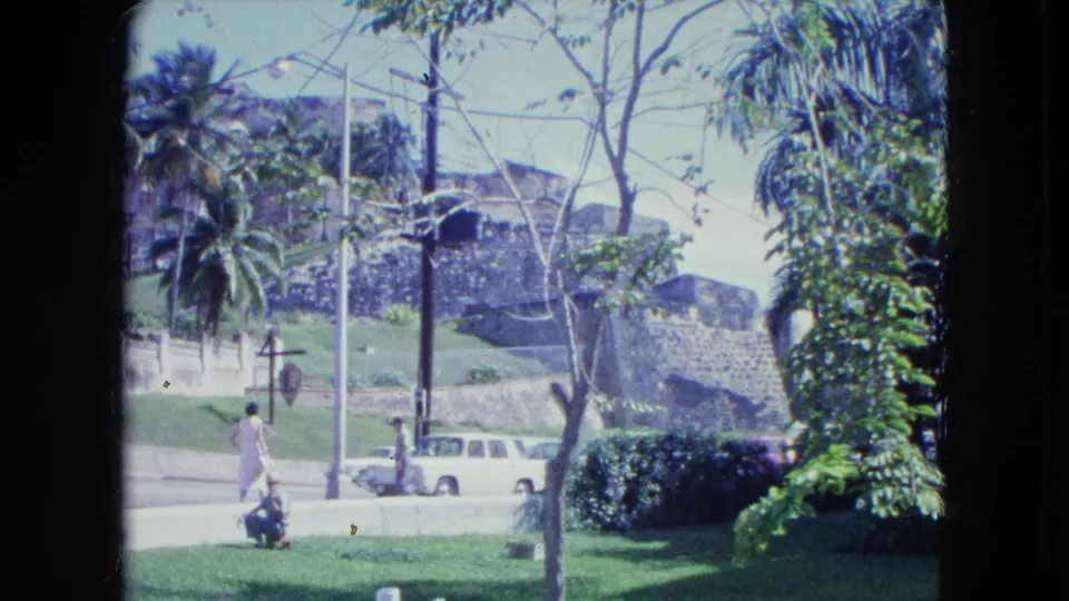 San Juan 1964 home-movie frame showing a pedestrian near the curb, a white station wagon, palms, and stone architecture.