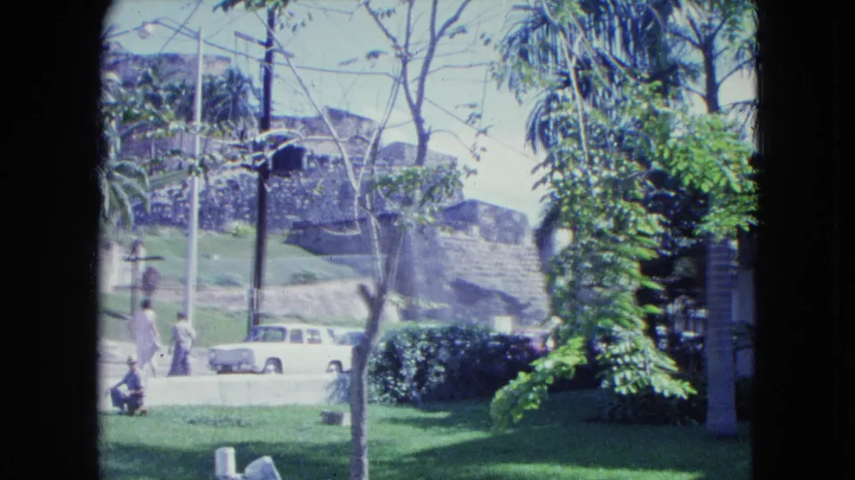 1964 San Juan street view with two pedestrians, a white station wagon, hedges, palms, and historic stone walls in the background.