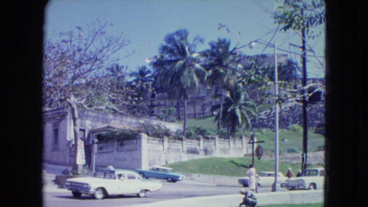 1964 San Juan street view with classic cars passing below stone fortification walls, palm trees, pedestrians, and utility poles.