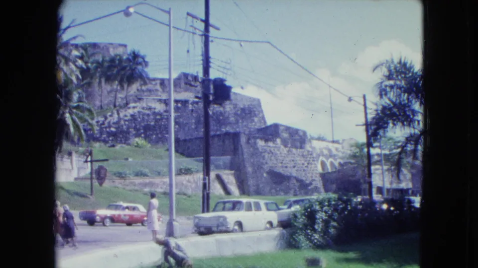 1964 San Juan street view with a red car, white car, pedestrians, palm trees, garden planting, utility poles, and stone walls.