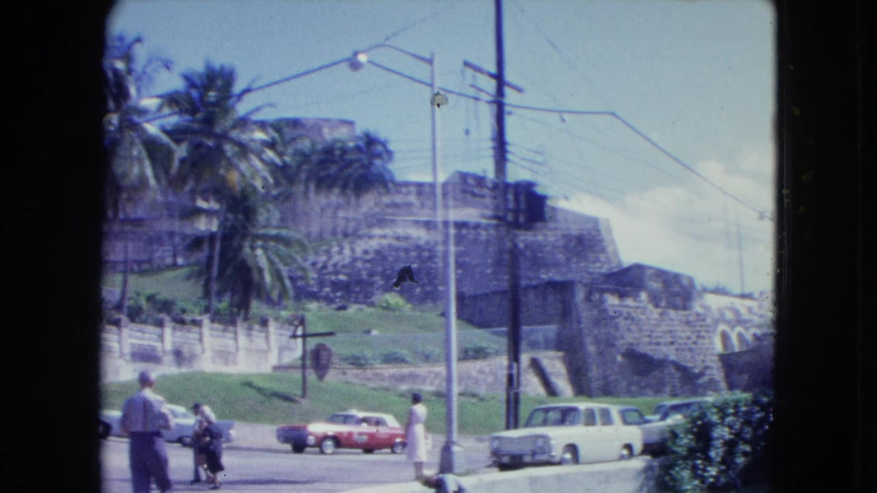 Pedestrians and cars pass below stone fortress walls with palm trees and streetlights.