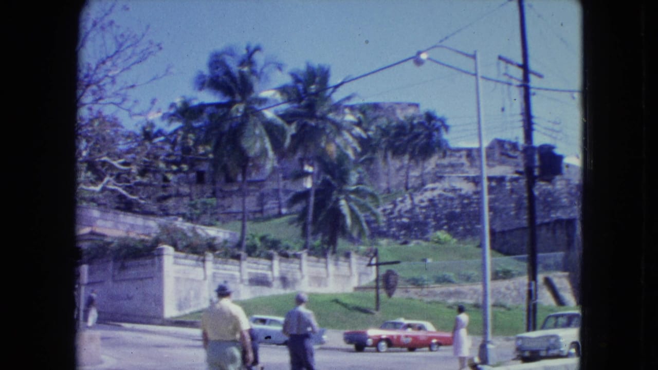 People walk near a roadside with classic cars and a stone fortification behind them.