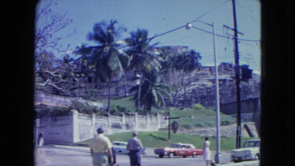 Street-level 1964 San Juan view of pedestrians walking past classic cars near palm trees and fortress masonry.