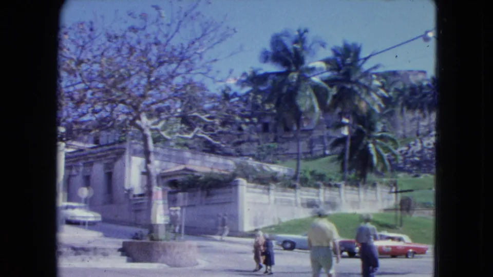 1964 San Juan roadside scene with walkers crossing below palm trees and the stone fortress structure.