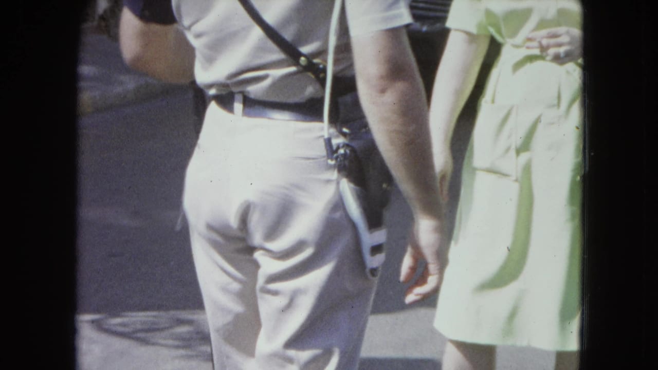 1964 San Juan home-movie frame showing a police officer from behind near a woman in a pale green dress on a bright street.
