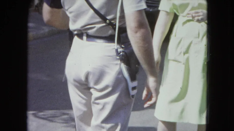 1964 San Juan home-movie frame showing a police officer from behind near a woman in a pale green dress on a bright street.