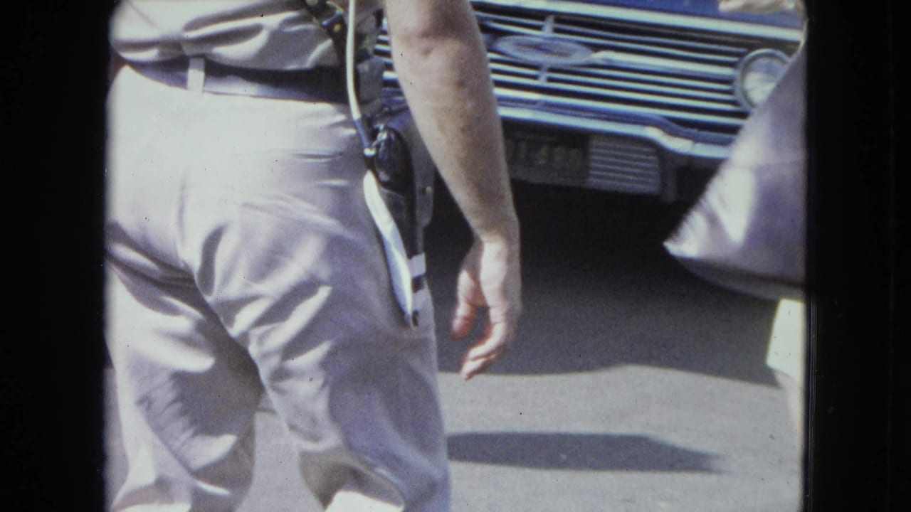 Police officer seen from behind walking near the front of a classic car and passing pedestrians.