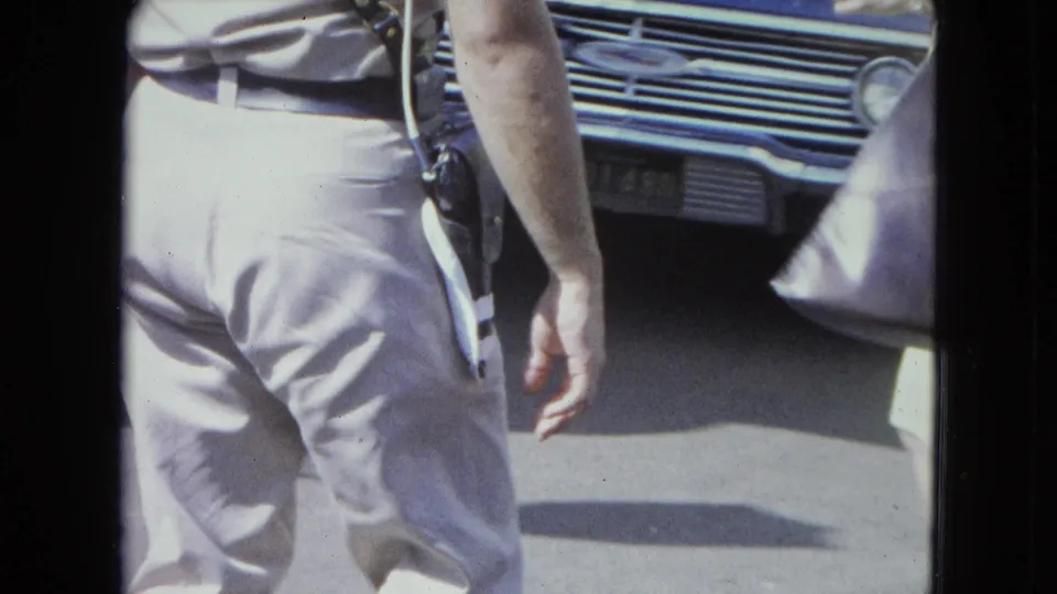 1964 San Juan street frame with a uniformed police officer from behind near the grille of a classic car and pedestrians.