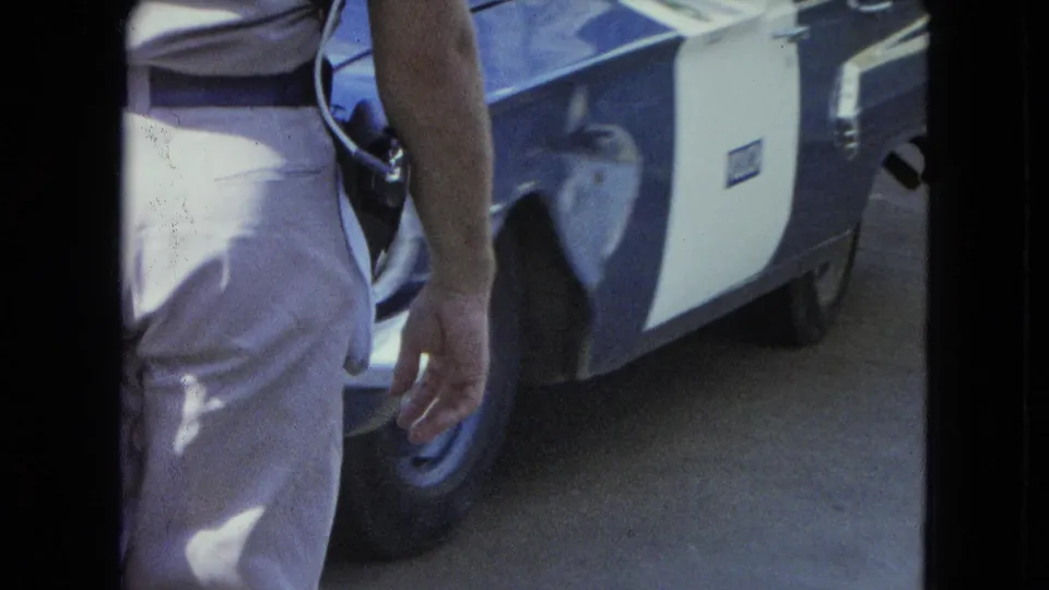 1964 San Juan home-movie frame showing the side of a uniformed officer with a holstered gun beside a blue and white car.