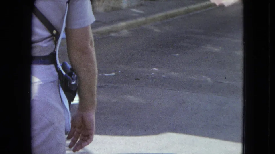 1964 San Juan frame with a partial rear view of a uniformed police officer near pavement and a pedestrian at frame edge.