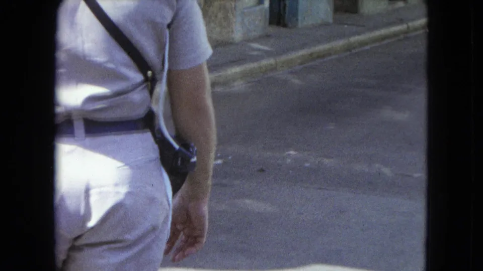 Light-uniformed man with belt, shoulder strap, and side-mounted radio walking along a San Juan street in 1964 footage.