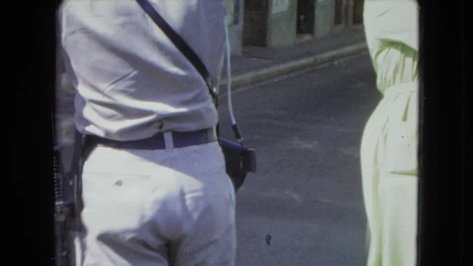 1964 San Juan frame showing a uniformed man from behind with a side radio and a partial passerby at the edge.