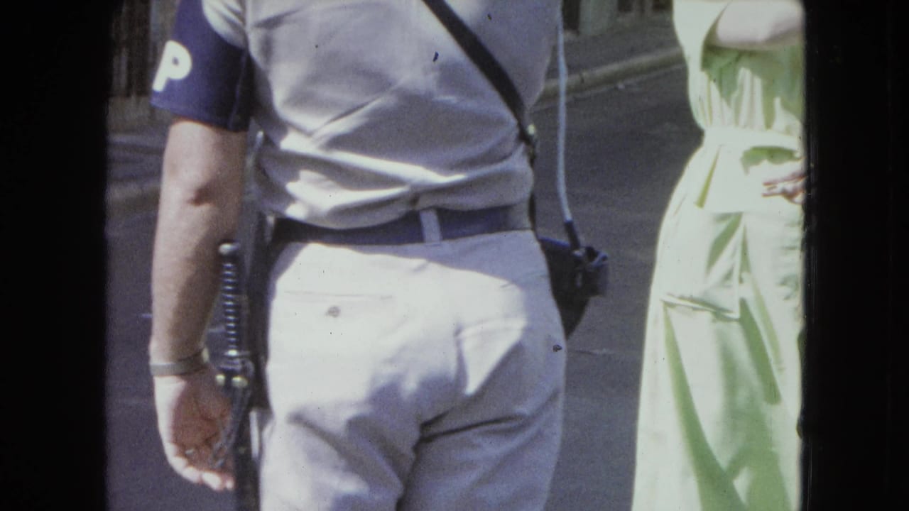Officer's back and sleeve armband are visible as he walks near a woman in green.