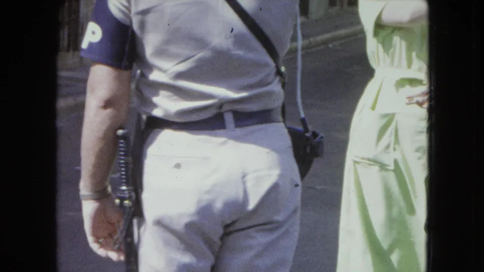 1964 San Juan frame showing the officer's back, blue armband with a white P, baton, sidearm, and a woman in pale green at frame edge.