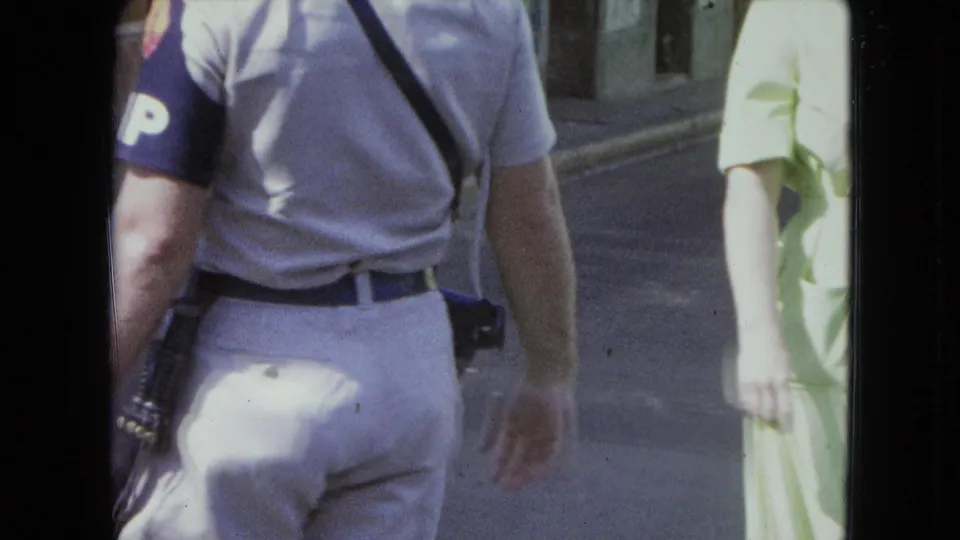 1964 San Juan street footage frames the officer from behind with baton and sidearm while a woman in a pale green dress passes at right.