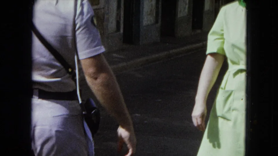 1964 San Juan frame with the officer partly turned away, shoulder strap across his back, and a woman in pale green walking at right.
