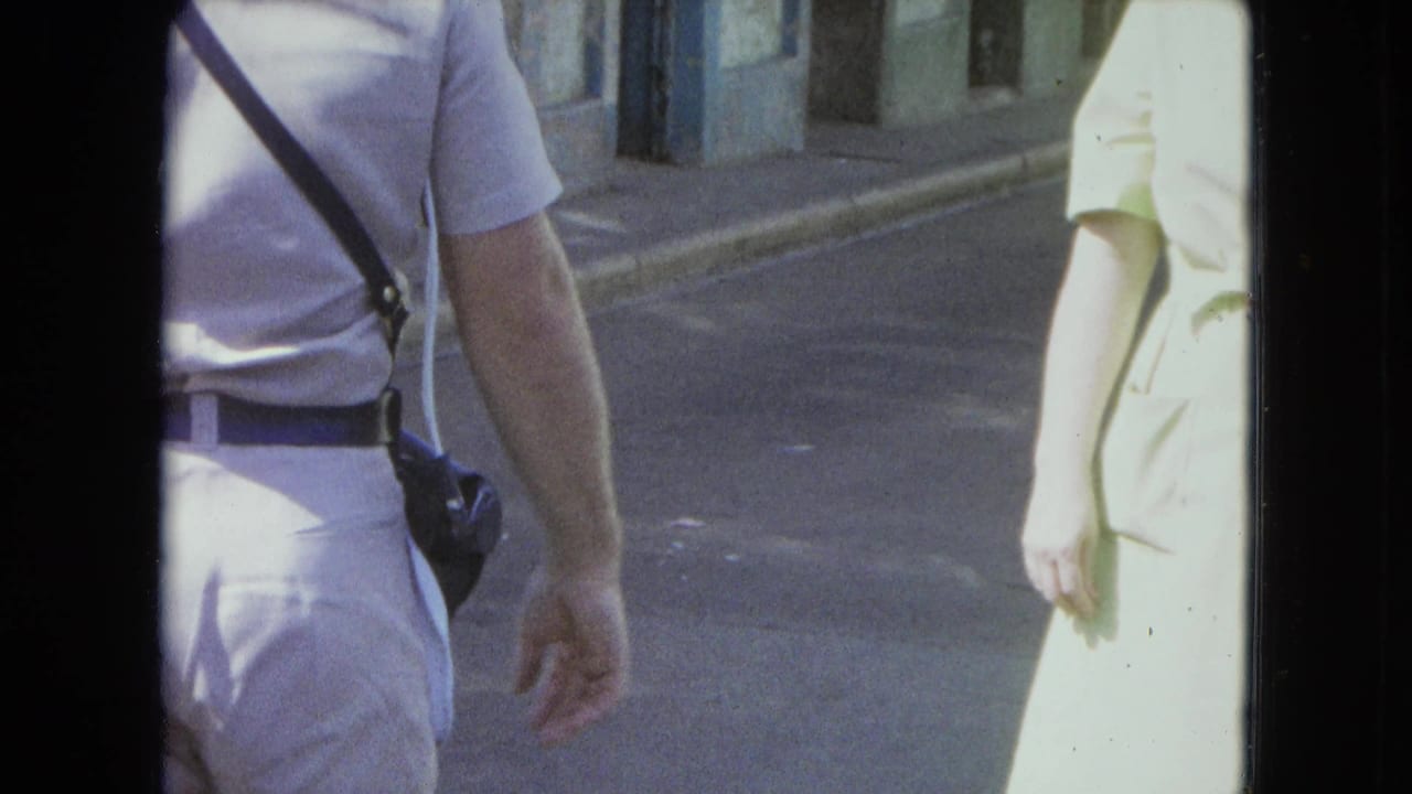 Man with camera strap and woman in pale dress walking on a sunlit street.