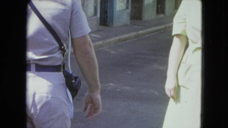 1964 San Juan home-movie frame showing a man with a camera strap beside a woman on a sunlit street.