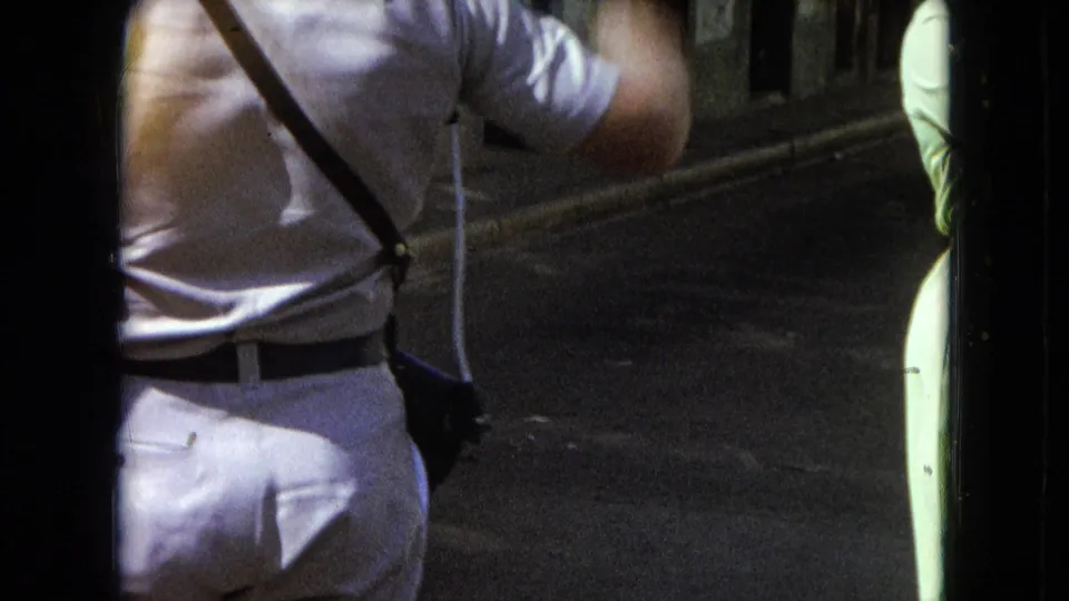 1964 San Juan frame showing a man lifting his arm as if waving while a woman stands near the street edge.