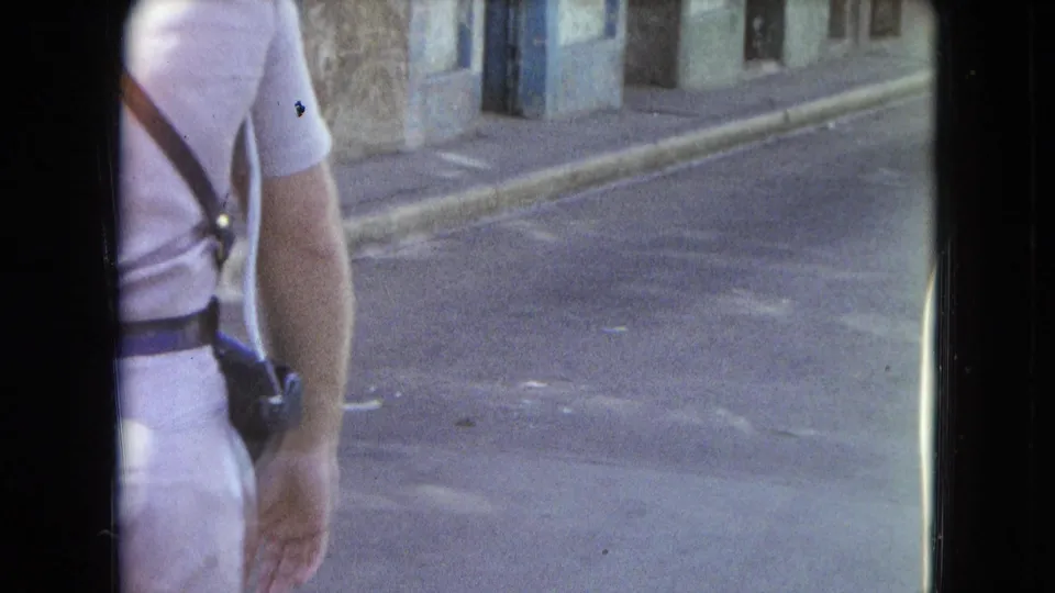 1964 San Juan home-movie frame showing a partial adult figure walking past a quiet street with aged building fronts.