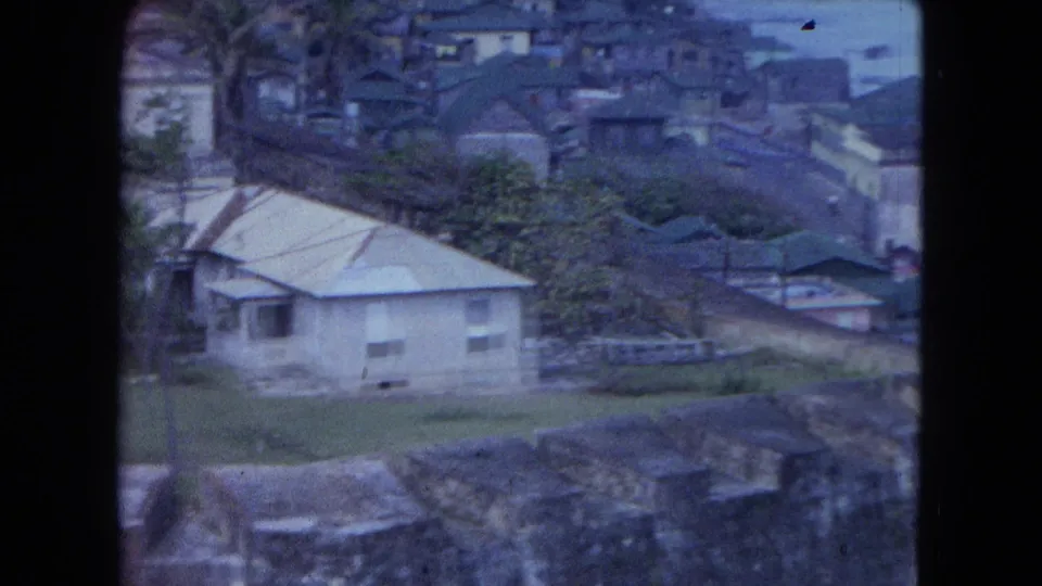 1964 San Juan home-movie frame showing a pale low building, trees, rooftops, and distant water beyond a hillside neighborhood.