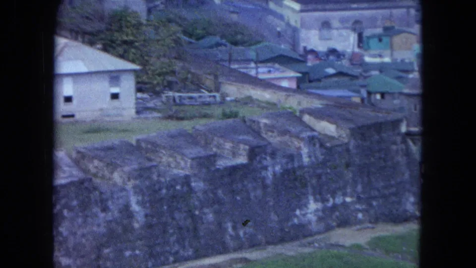1964 San Juan view with a weathered fortress wall in the foreground, low buildings behind it, and the coastline visible beyond.