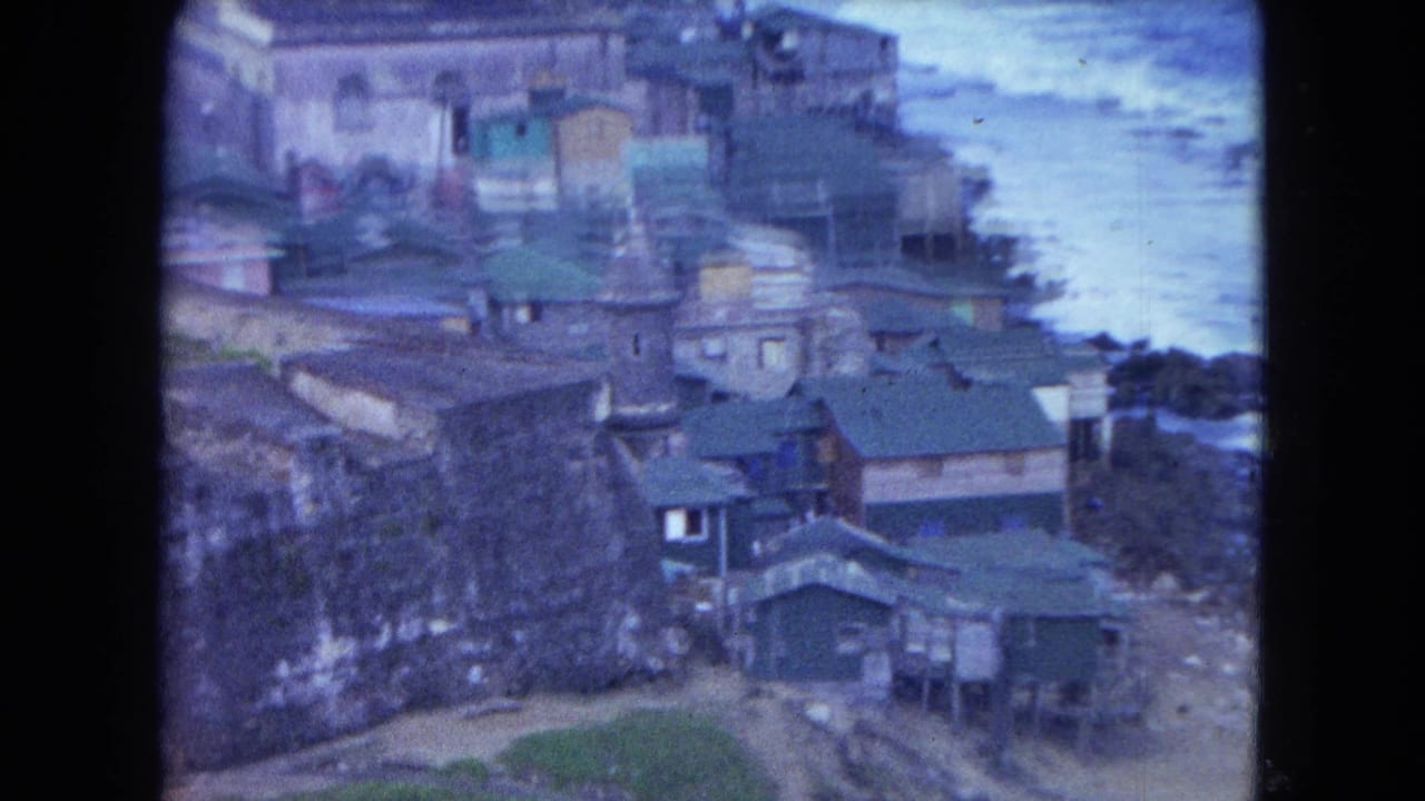 1964 San Juan shoreline view with houses, a high stone wall, green roofs, and surf at the right edge.