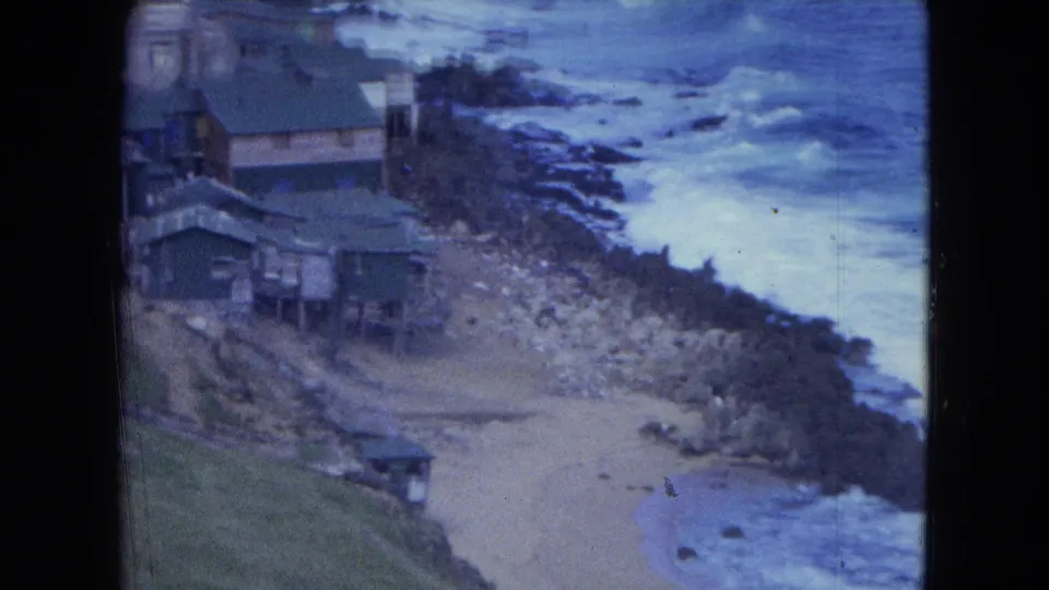 1964 San Juan coastal frame showing stilted houses, a small beach, rocks, and waves along the shore.