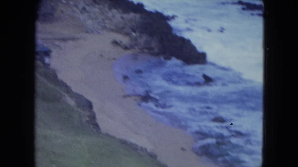 1964 San Juan shoreline view focused on a sandy beach, rocks, grass, and waves breaking near shore.