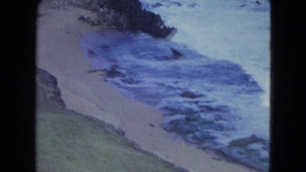 1964 San Juan beach view on 8mm, showing foamy surf, dark rocks, sand, and a grassy coastal slope from above.