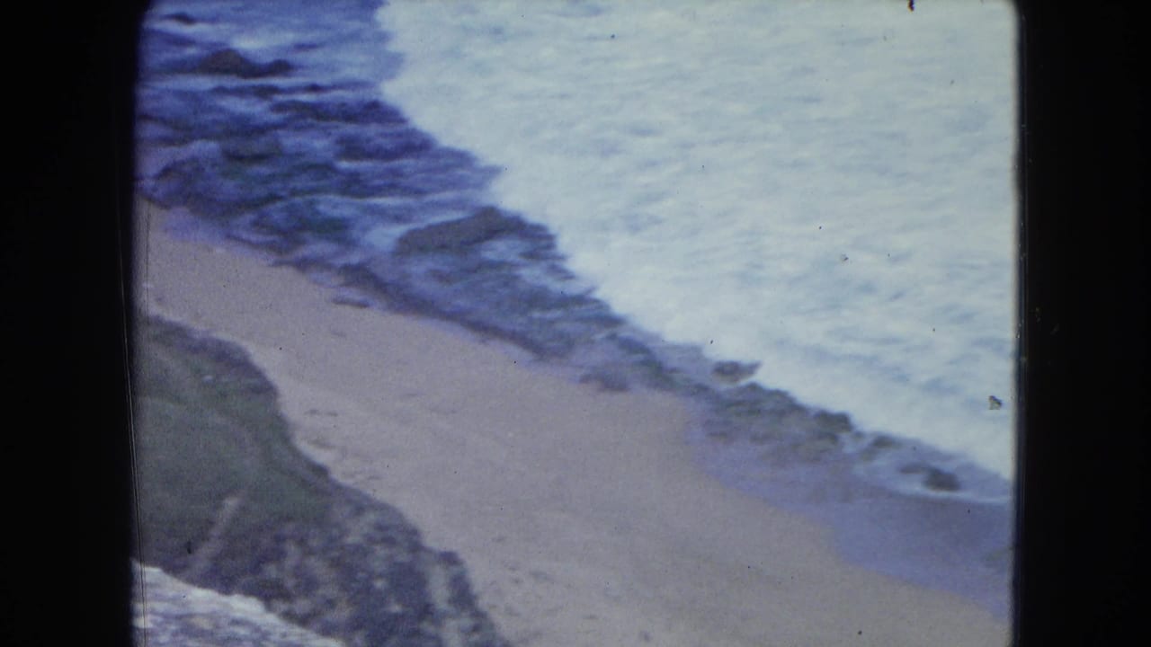 1964 San Juan coastal frame with ocean surf cutting across a sandy beach below rocks and grass-covered shoreline.