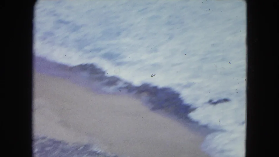 Soft 8mm view of San Juan shoreline, with pale surf rolling over a narrow beach and scattered dark rocks.