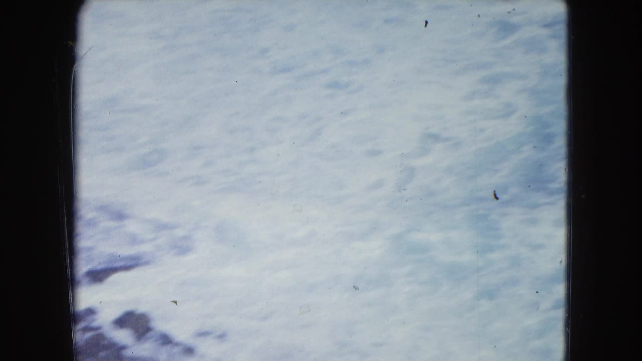 1964 San Juan shoreline footage shows pale blue water and white foam seen close up through visible 8mm gate edges.