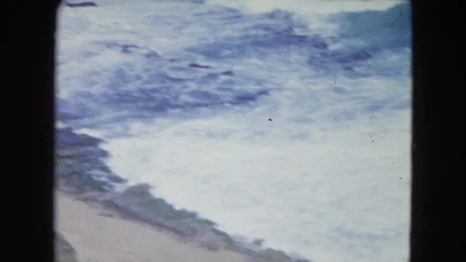 1964 San Juan beach footage shows foamy ocean waves reaching pale sand beside darker rocks, with visible film grain and fading.