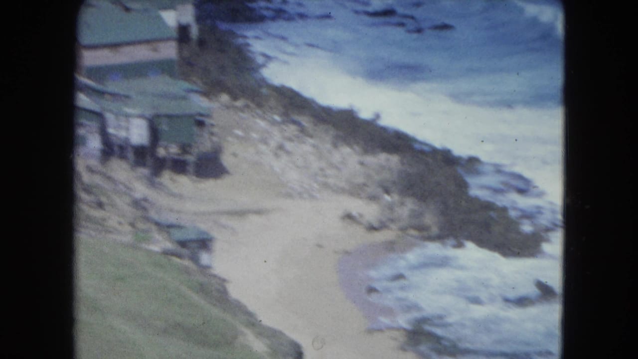 Rocky coastline with surf, sandy beach, and small buildings on a hillside.