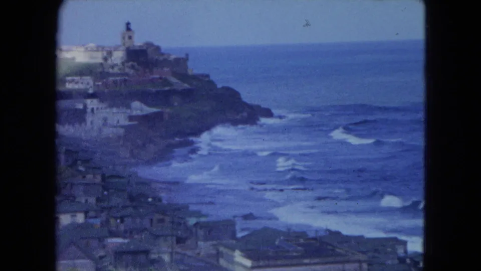1964 8mm frame of a San Juan coastal hillside, with a tower-like structure, rooftops, open sea, and surf below.
