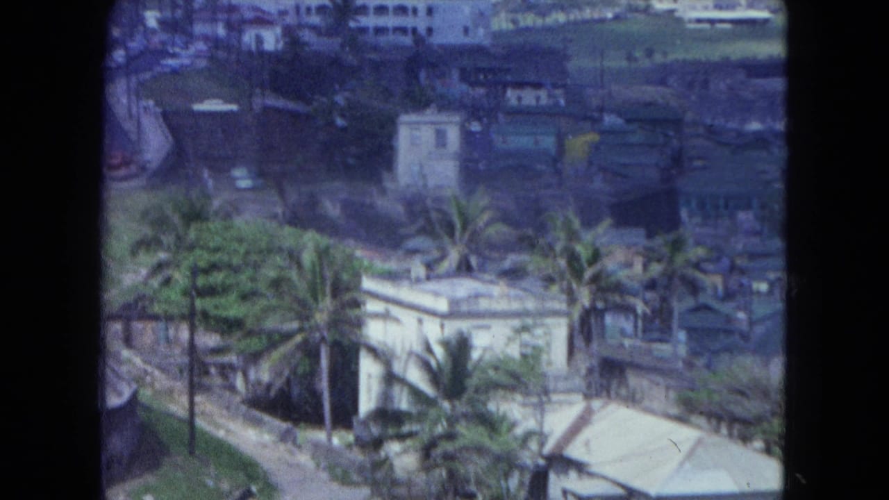 1964 San Juan home-movie frame showing a hillside residential area with palms, white buildings, and a winding road.