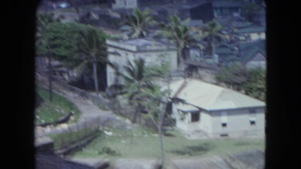 1964 San Juan frame with a white house, palm trees, grass, and a road in a bright residential hillside view.