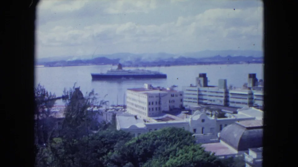 1964 San Juan harbor view with a large ocean liner, waterfront buildings, open water, and distant mountains.