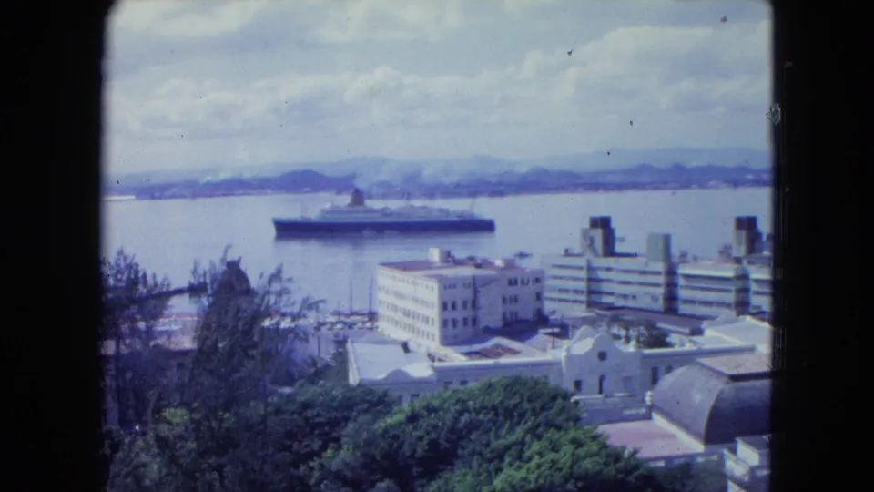 1964 San Juan harbor view from above, showing waterfront buildings, trees, distant hills, and an ocean liner offshore.