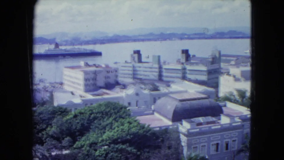1964 San Juan shoreline from above, showing waterfront architecture, a domed roof, trees, harbor water, and a partial ocean liner.