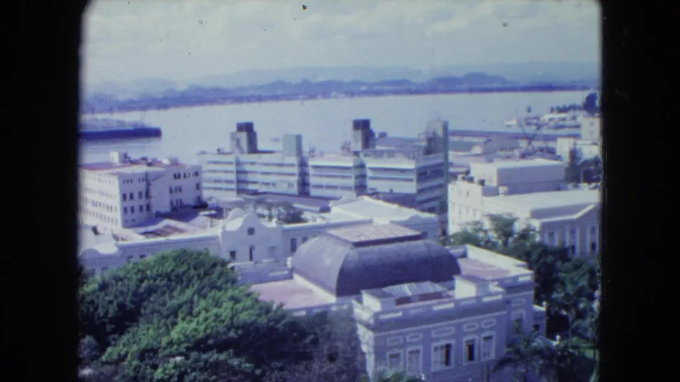 1964 San Juan harbor view with foreground trees, waterfront buildings, a partial ship at left, and distant hills across the water.