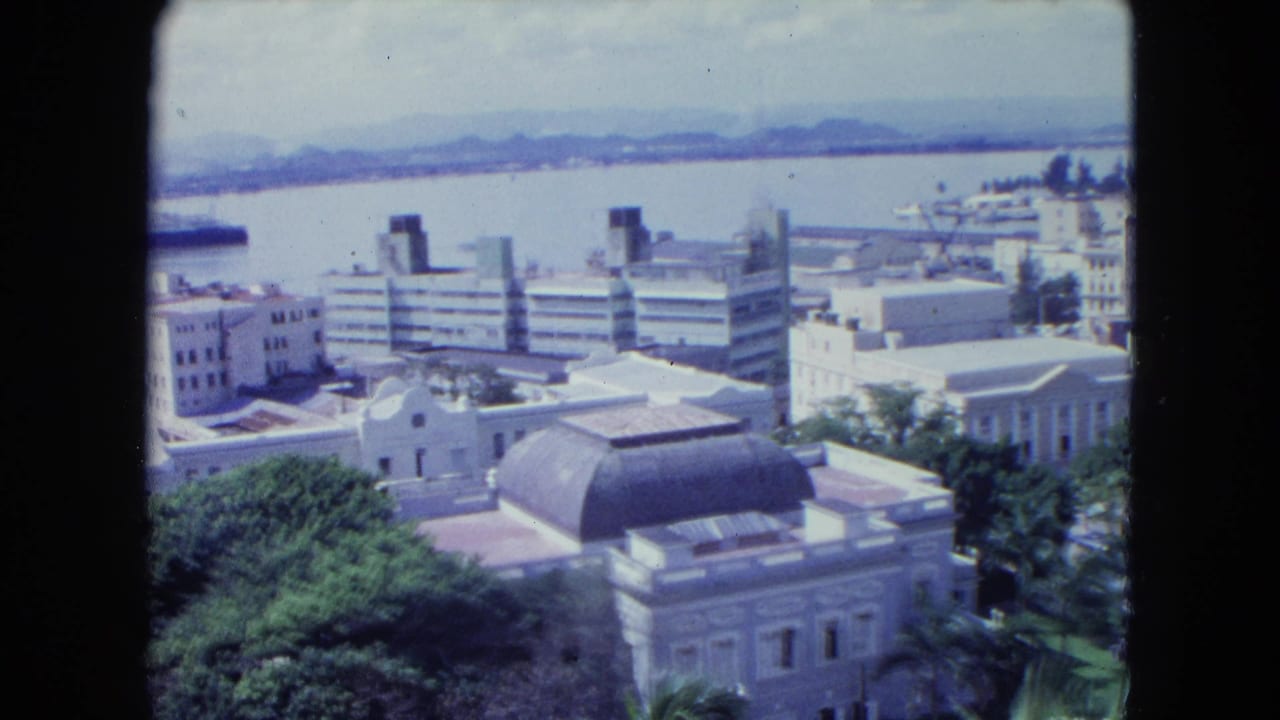 1964 San Juan rooftop view showing a domed building, waterfront blocks, trees, bright water, and hazy hills beyond the city.
