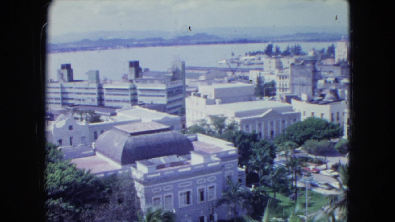 Elevated 1964 San Juan view with downtown rooftops, a domed building, roadside greenery, waterfront, and hazy hills.