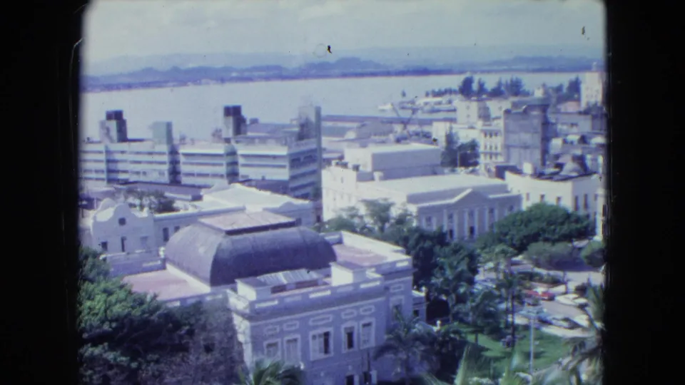 Elevated 1964 San Juan view with downtown rooftops, a domed building, roadside greenery, waterfront, and hazy hills.